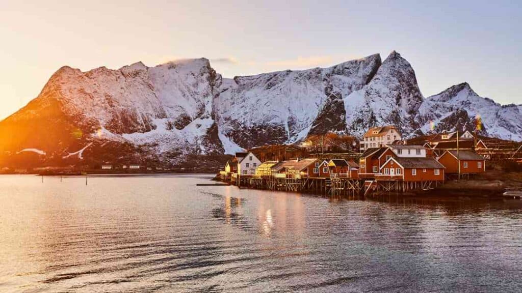 A small coastal village with red and white wooden houses by the sea in front of massive snow covered Arctic mountains in a golden winter sun. Find out how to get to Lofoten here. 