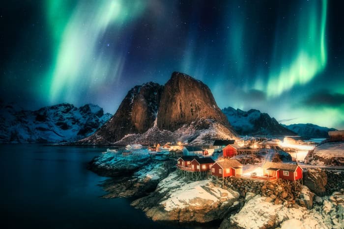 Dancing green northern lights on a dark sky over a small fishing village with red cabins next to the fjord and vast majestic mountains