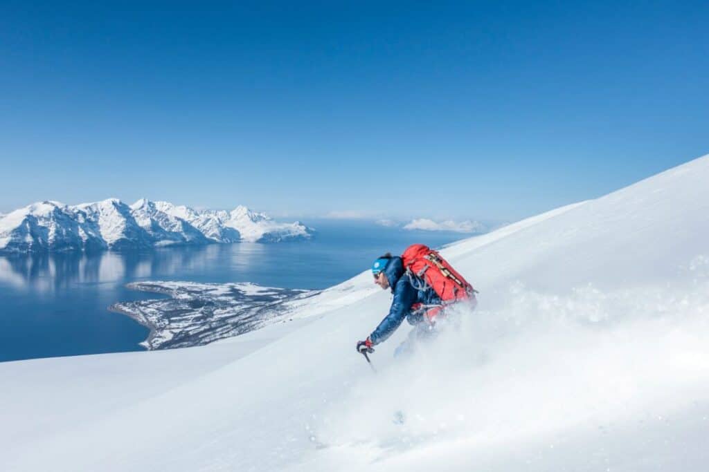 An off-pist skier going down a mountain in bright sunshine with a backdrop of a dark blue blank sea and rugged mountains covered in snow