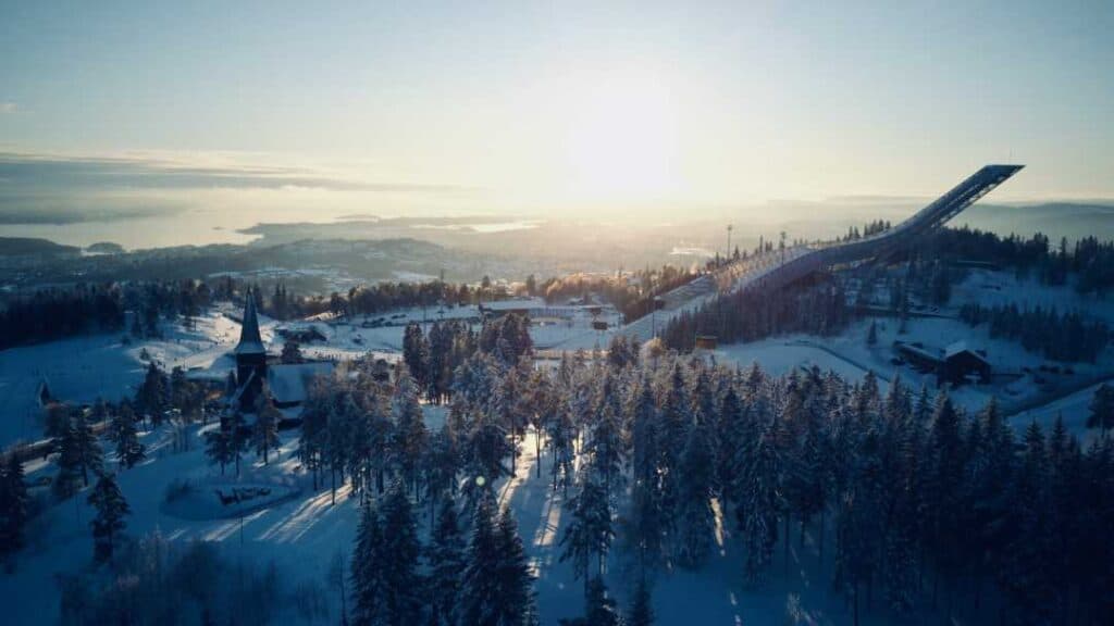 Birdseye view of a forest and a huge ski jump in winter, the ground covered in snow under a pale blue sky with a low winter sun