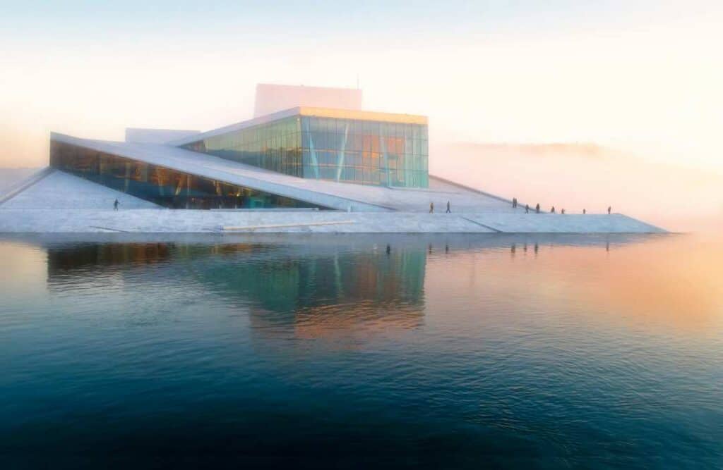 A white modernistic stone and glass building sitting on the dark water surrounded by white mist on a winter day, magically lit by the sun behnind the mist creating a glow in the photo