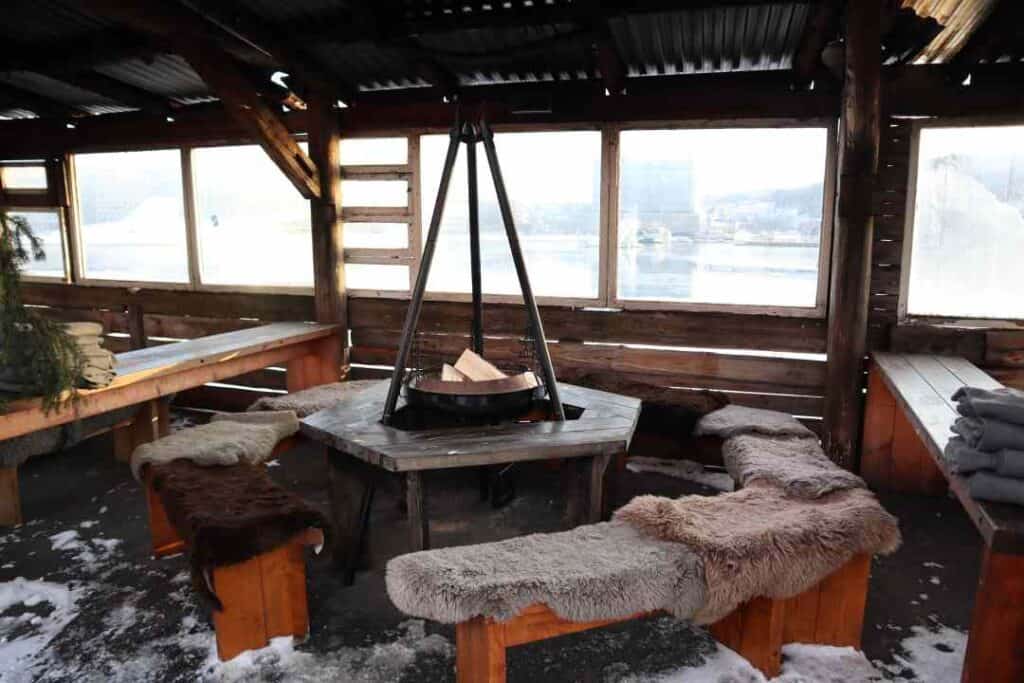 A table with a bonfire in the middle, with benches around it covered with furs for restaurant seating in winter with fjord views through the windows