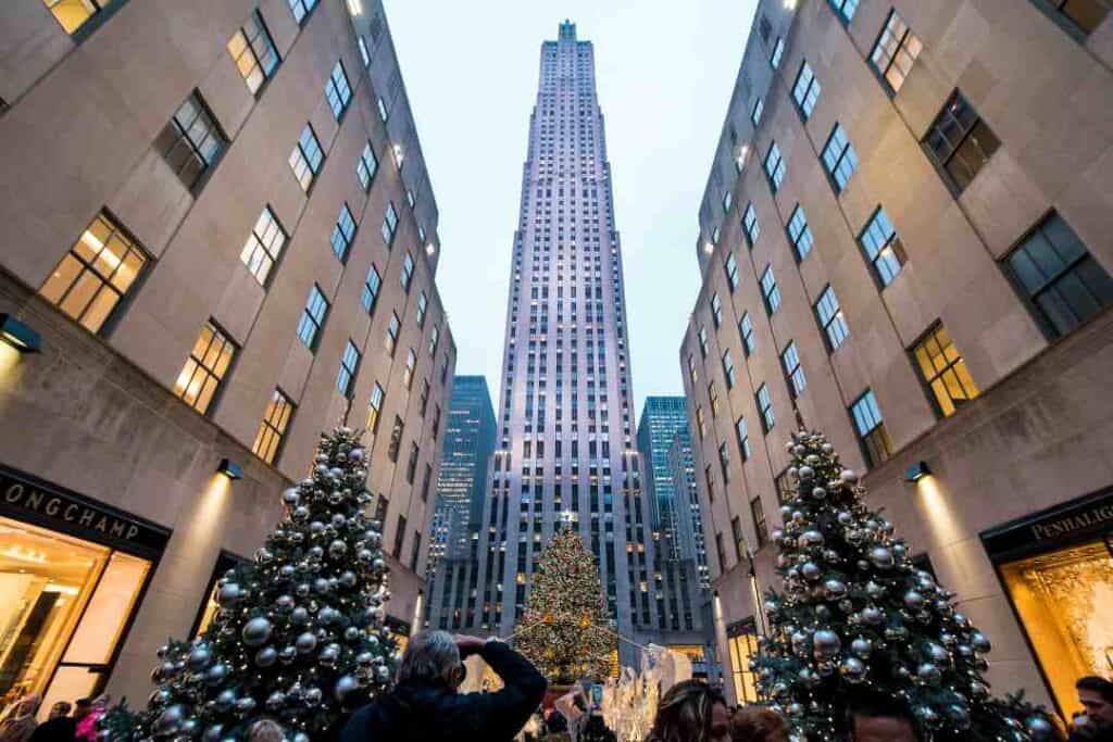 a tall skyscraper stretching to the sky between two other modern stone buildings, and in the fron are decorated christmas trees and people admiring the view