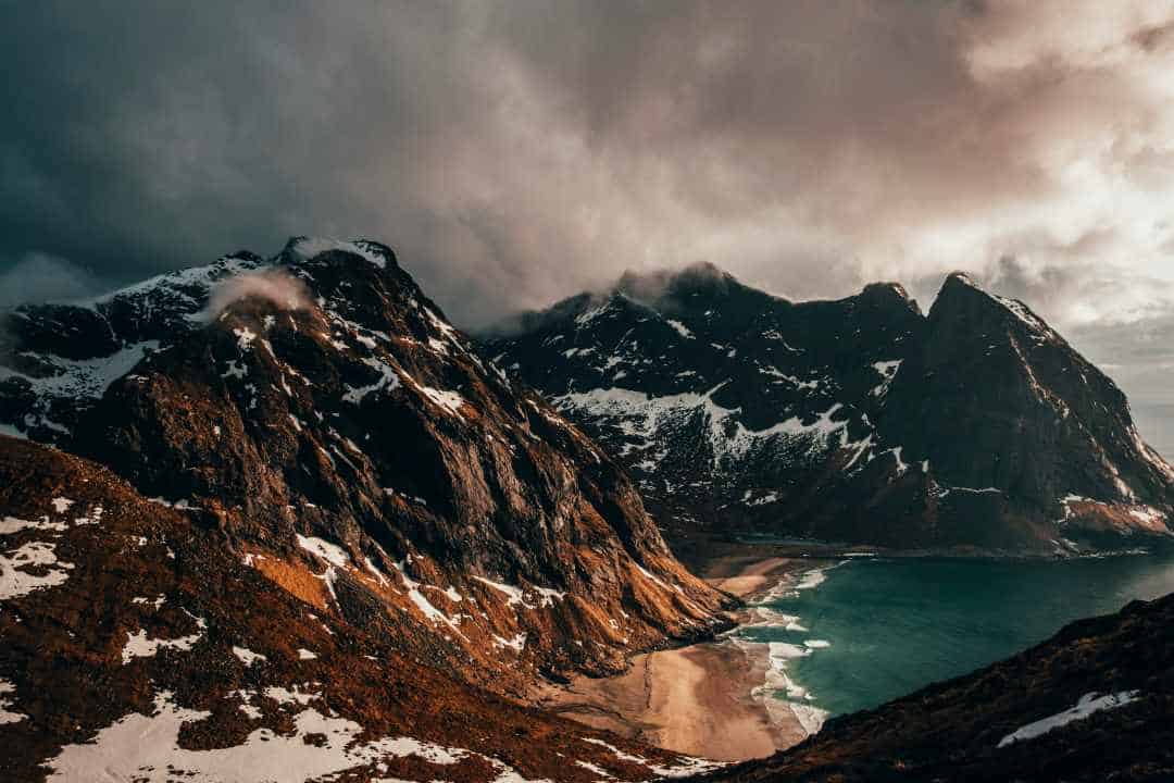 A massive mountain range with dramatic drops down to a sandy beach under dark clouds, yet in golden light from a low sun in the distance