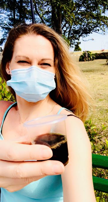 Woman with long blond hair in a park, smiling behind a face mask, holding a small cup of strong, dark coffee 