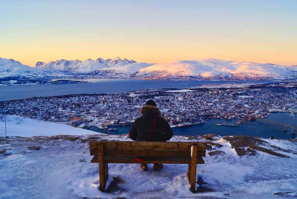 A man sitting on a wooden bench in winter on top of a mountain looking down on a town on an island below with slow covred mountains in the background