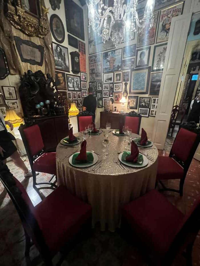 A beautifully set round dinner table surrounded by red velvet chairs, a golden table cloth, and the interior around is full of photos, art, and stunning details undet the high ceiling