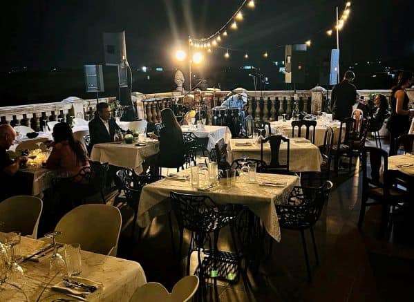 An outdoor rooftop restaurant at night, with warm lighting, white table cloths, and people enjoying themselves with dinner