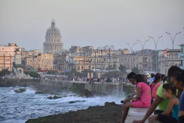 a city boardwalk at sunset with strong waves hitting the wall, and lots of people relaxing on the brick wall of the boardwalk with a rugged city skyline in the background